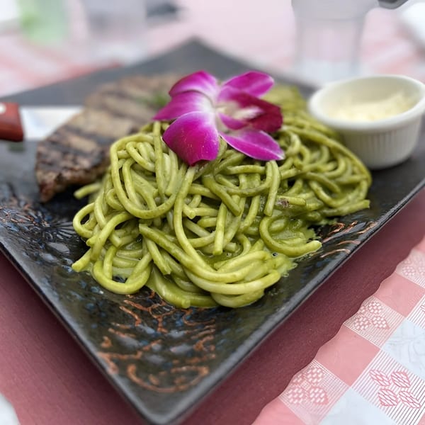 Peruvian green spaghetti with grilled steak and a purple orchid garnish on a dark plate.